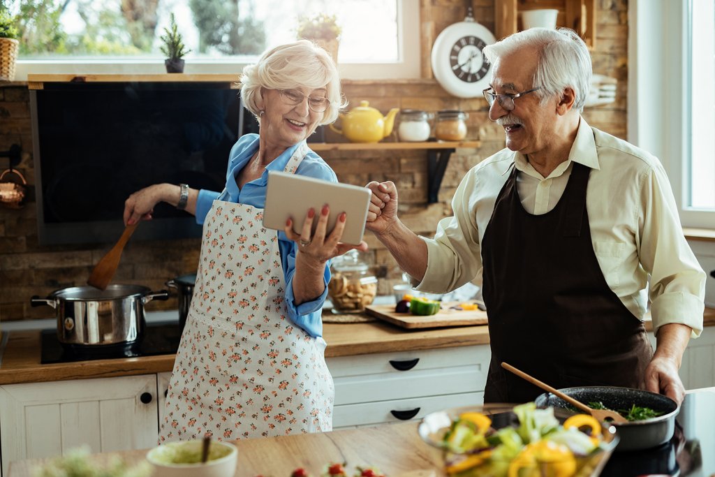 Happy old couple cooking in the kitchen. Cheerful, senior, husband and wife couple having fun while preparing food, using a tablet, in their kitchen. Image by Drazen Zigic on Freepik.