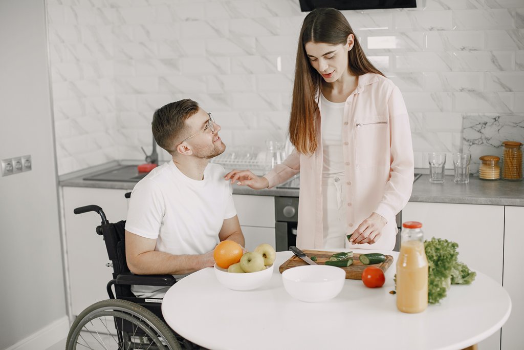 Woman and man in a wheelchair sat at kitchen table, preparing food in the kitchen.
