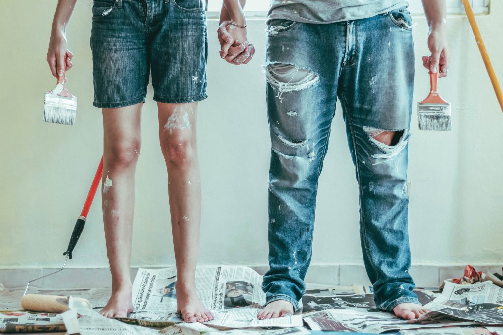 Man and woman's legs, barefoot, wearing jeans and splattered with paint from DIY painting and decorating. Roselyn Tirado from Unsplash.